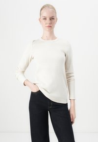 Lange mouw, crèmekleurige geribbelde top met een ronde hals, gecombineerd met donkere spijkerbroek. Minimalistisch ontwerp, zachte textuur en een aansluitende silhoeet.