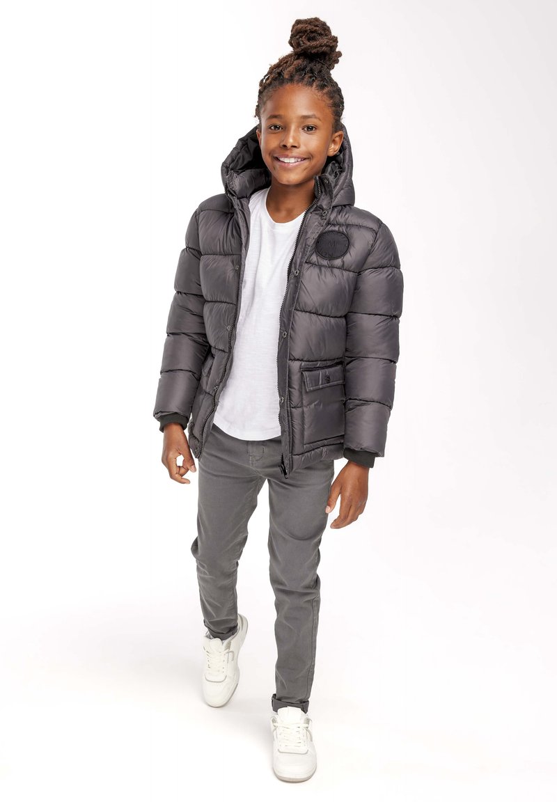 Black puffer jacket with a hood, featuring quilted design, a front zipper, and a patch on the chest. Paired with gray pants and white sneakers.