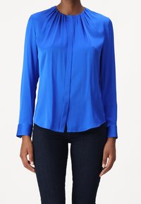 Kvinna som bär en långärmad, ljusblå blus med samlad halsringning och mörkblå tajta jeans, stående mot en enkel bakgrund.