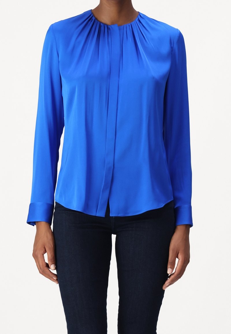 Kvinna som bär en långärmad, ljusblå blus med samlad halsringning och mörkblå tajta jeans, stående mot en enkel bakgrund.