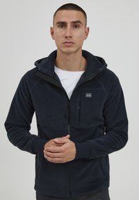 Marinefarget fleece zip-up hettegenser med frontlomme, snøring i hetten og kontrastfarget detalj ved glidelåsen. Myk tekstur.