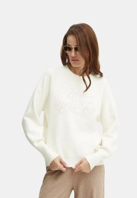 Felpa bianca con polsini e orlo a coste, con un logo "Guess" testurizzato sulla parte anteriore, indossata con pantaloni marrone chiaro.