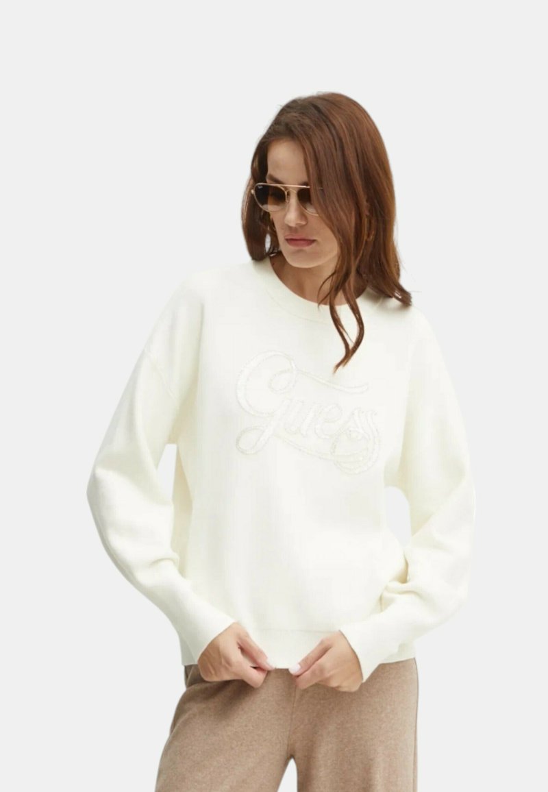 Felpa bianca con polsini e orlo a coste, con un logo "Guess" testurizzato sulla parte anteriore, indossata con pantaloni marrone chiaro.