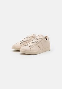 Ljusbeige lädersneakers med låg profil, rund tå, snörning och texturerade accenter på sidorna. Slät yta och minimalt mönster.