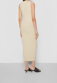 Beige pletené midi šaty s přiléhavým střihem a bezrukávovým designem, kombinované s černými otevřenými lodičkami na podpatku. Hladká textura.