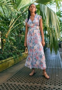 Gėlėta maxi suknelė šviesiai mėlynos spalvos, su rožinėmis ir žaliomis gėlėmis, prigludusi juosmens dalis su raukšlėtu apykakliu; derinama su auksinėmis sandalais ir rankine.