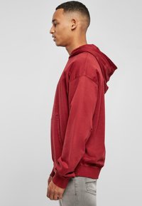 Giovane uomo in piedi di lato, che indossa un hoodie rosso usurato e jeans grigio chiaro, su uno sfondo grigio semplice.