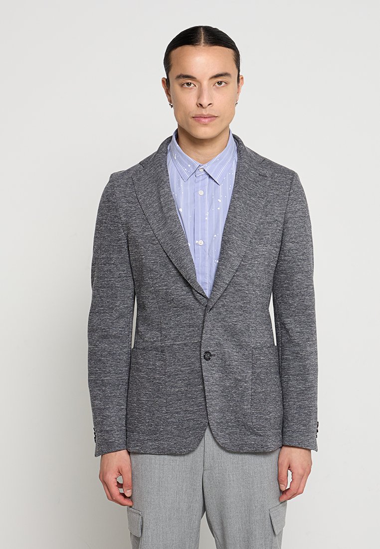 Szary, teksturowany blazer z klapami, zapięciem na jeden guzik i dwiema przednimi kieszeniami, noszony na jasnoniebieskiej koszuli w wzór.