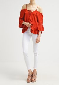 Blouse rouge décolletée avec un nœud à l'avant, manches évasées et tissu texturé, associée à un jean slim blanc et des talons nude.
