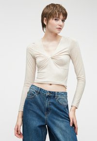Beige langærmet top med V-udskæring og samlet frontdetalje, parret med højtaljede blå jeans med et klassisk fem-lomme design.