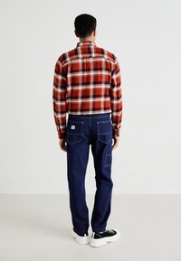 Personne portant une chemise à carreaux rouge et blanc, un jean bleu avec des poches à rabat, et des baskets noir et blanc, se tenant de dos face à l'appareil photo.