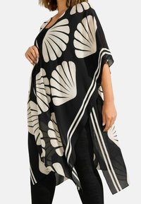 Musta läpinäkyvä kimono, jossa on valkoinen simpukkakuviot, leveät hihat ja raidalliset reunat, valuu löysästi tummien housujen yli.