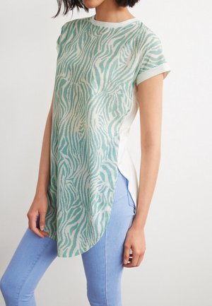 Femme portant une tunique à manches courtes, à motif zébré vert et blanc, avec des revers aux manches, et un jean bleu clair sur un fond uni.