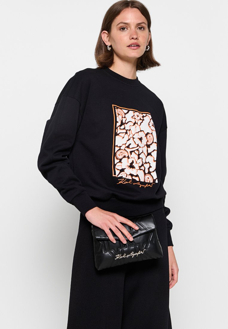 Felpa nera con stampa floreale bianca e arancione, caratterizzata da una vestibilità rilassata. Il modello tiene in mano una borsa a tracolla nera imbottita con testo dorato.
