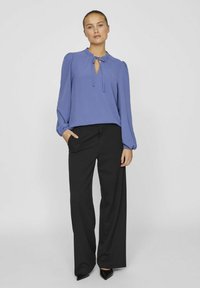 Blusa blu a maniche lunghe con scollo a chiave e dettaglio con lacci, abbinata a pantaloni neri a vita alta e gamba larga, e tacchi a punta neri.