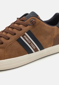 Sneaker en suède marron avec des rayures bleu marine et blanches, marqué "MARCO TOZZI," doté d'une semelle texturée et d'un design à laçage.
