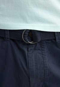 Camisa azul claro metida dentro de pantalones azul oscuro con un cinturón tejido azul oscuro asegurado por anillas dobles de metal.
