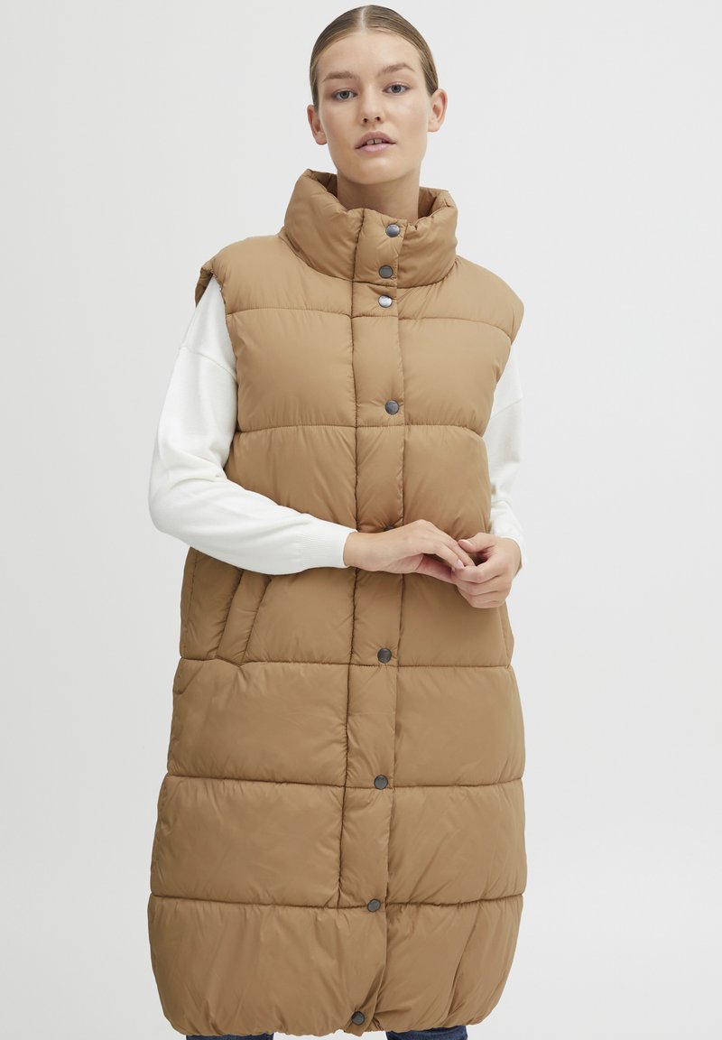 Brun polstret vest med høy krage, med store quiltede seksjoner og trykknapper. Brukt over en hvit langermet topp.