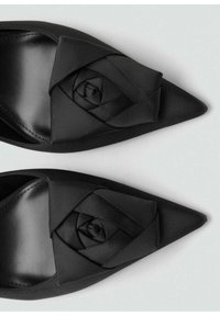 Zapatos de satén negros con punta afilada que presentan un diseño floral en 3D en la parte frontal. Textura suave con una forma estilizada. Sin herrajes ni acentos visibles.