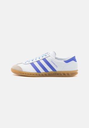 Zapatilla blanca con rayas moradas, puntera de ante y cordones azul claro. Suela de goma con marca "Adidas" en el lado.