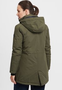 Olive green parka with a hood, featuring a zip closure, side pockets, and a slightly longer back hem; smooth texture with subtle stitching details.