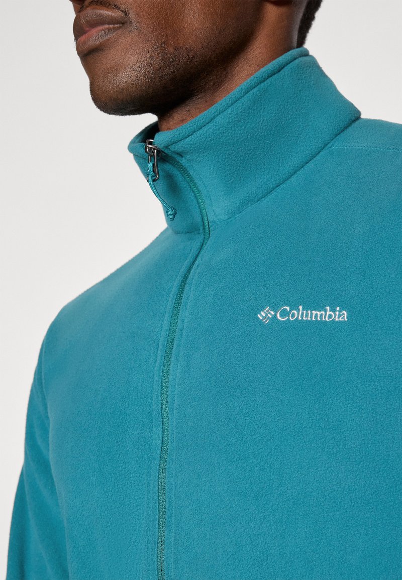 Zielona polarowa kurtka z wysokim kołnierzem, zamkiem z przodu oraz haftowanym logo "Columbia" na piersi. Miękka tekstura i gładkie wykończenie.