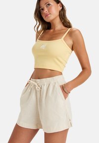 Haut court côtelé jaune avec des fines bretelles et un petit logo sur le devant, associé à un short en lin beige avec une taille élastique et des poches.