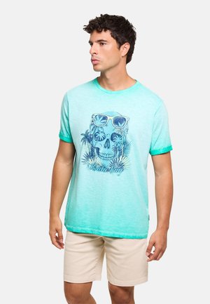 Hombre que lleva una camiseta turquesa claro con diseño de calavera, palmeras y gafas de sol, combinado con pantalones cortos beige, mirando hacia un lado.