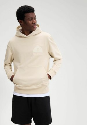 Mann trägt beige Kapuzenpullover mit Vordertasche und schwarze Shorts, schaut seitlich vor einem schlichten hellen Hintergrund.
