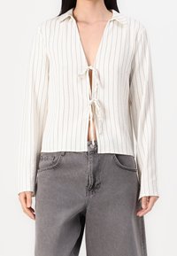 Femme portant un haut crème à manches longues avec des rayures verticales et des fermetures à nouer à l'avant, associé à un jean décontracté en denim gris.