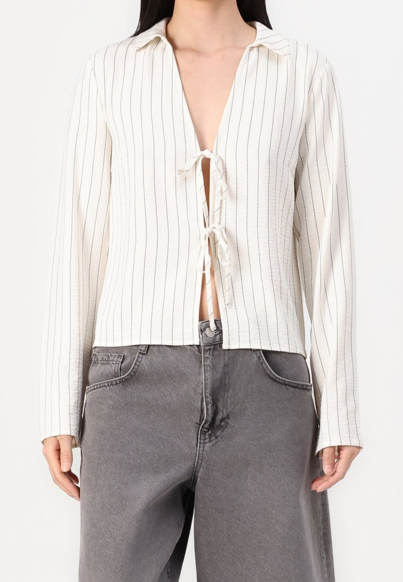 Femme portant un haut crème à manches longues avec des rayures verticales et des fermetures à nouer à l'avant, associé à un jean décontracté en denim gris.