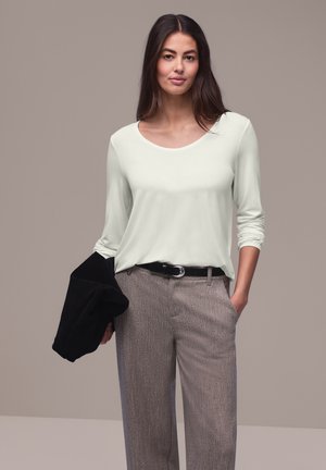 Mujer de cabello largo y oscuro que lleva una camisa de manga larga color crema, pantalones grises con cinturón negro, sosteniendo una chaqueta negra doblada en una mano.