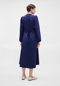 Sötétkék ruha függőleges csíkokkal, hosszú ujjal és megkötős derékkal. Simább anyag, midi hosszúságú és egyenes szabású.