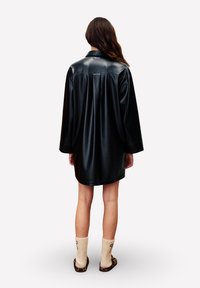 Veste-chemise surdimensionnée en cuir noir avec col, manches longues et coupe ample. Portée avec des chaussettes beiges et des chaussures à enfiler à imprimé animal.