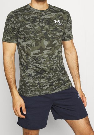 Ein Mann trägt ein grünes Digital-Camouflage-T-Shirt von Under Armour und dunkelblaue Adidas-Sportshorts und steht vor weißem Hintergrund.