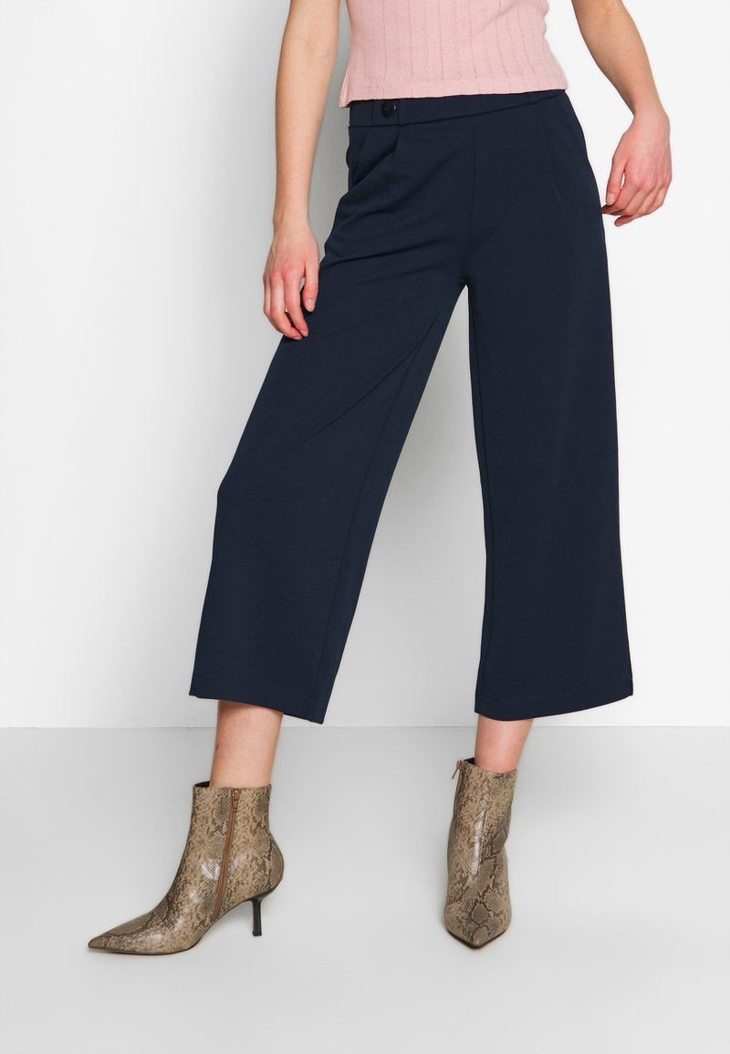 Pantalons larges bleu marine avec une taille haute et des plis à l'avant, associés à des bottines à talons imitant un serpent brun, avec un embout pointu et des zips.