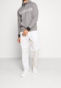 Sweat à capuche gris avec le texte "CALVIN KLEIN" en blanc, associé à un pantalon blanc à rayures et des baskets blanches. Design simple et décontracté.