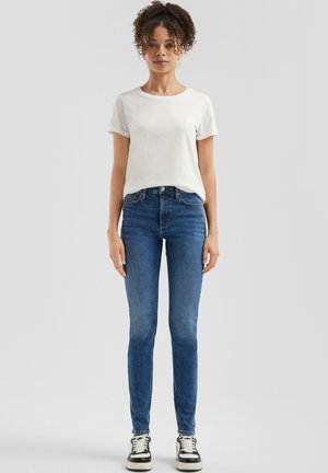 Weißes T-Shirt, kurze Ärmel, kombiniert mit blauen Skinny-Jeans und schwarz-weißen Sneakers. Model steht frontale mit neutralem Hintergrund.