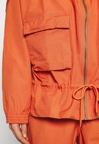 Orange lättviktig jacka med elastiska ärmslut och dragsko i midjan. Har en dragkedjestängning och två stora framfickor med lappar. Texturerad tyg.