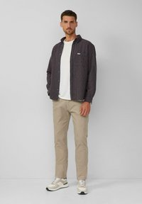 Man in beige broek, witte sneakers, wit T-shirt en open donker geblokt overhemd staand tegen een effen lichte achtergrond.