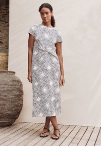 Jurk met korte mouwen van een lichte stof met een geometrisch zwart-wit patroon, knoopdetail in de taille en een lengte tot aan de enkels.