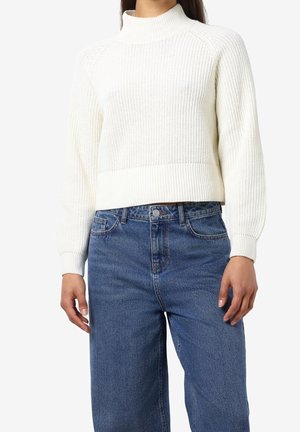 Personne portant un pull blanc en tricot à manches longues et un jean bleu taille haute sur un fond uni.