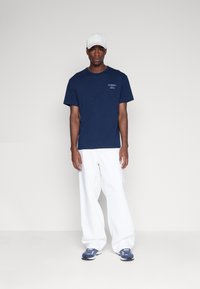 Camiseta de algodón azul marino con el logo "TOMMY JEANS" en el pecho, combinada con pantalones holgados blancos y zapatillas deportivas azules. Gorro de béisbol blanco.