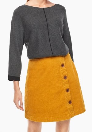 Kobieta ubrana w ciemnoszary top z długim rękawem oraz musztardowożółtą spódnicę sztruksową z czterema brązowymi guzikami z przodu po prawej stronie.