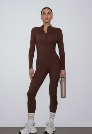 Kobieta nosząca dopasowany brązowy kombinezon, białe tenisówki, paskowane skarpetki, trzymająca beżową butelkę na wodę, stoi przy gładkiej ścianie.