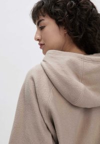 Felpa con cappuccio beige in tessuto testurizzato, dettagli di cucitura visibili e una vestibilità rilassata, che mostra il profilo di una donna di spalle.
