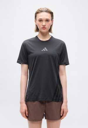 Junge Person mit kurzen blonden Haaren trägt ein schwarzes Adidas-T-Shirt und braune Shorts und steht vor einem schlichten weißen Hintergrund.