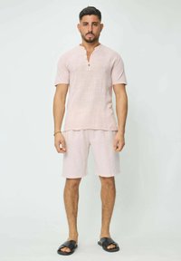 Chemise en lin rose pâle à manches courtes avec un col étroit et des boutons, associée à un short assorti ; des sandales noires à glissière complètent la tenue.