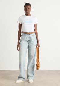 Hvit t-skjorte med blå grafisk tekst, lyseblå høytlivte jeans, lys brun lærbelte og hvite joggesko mot en nøytral bakgrunn.