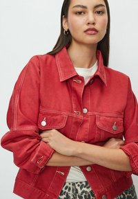 Young woman with long dark hair wearing gold hoop earrings, a red jacket with white stitching, a white shirt, and leopard print pants.